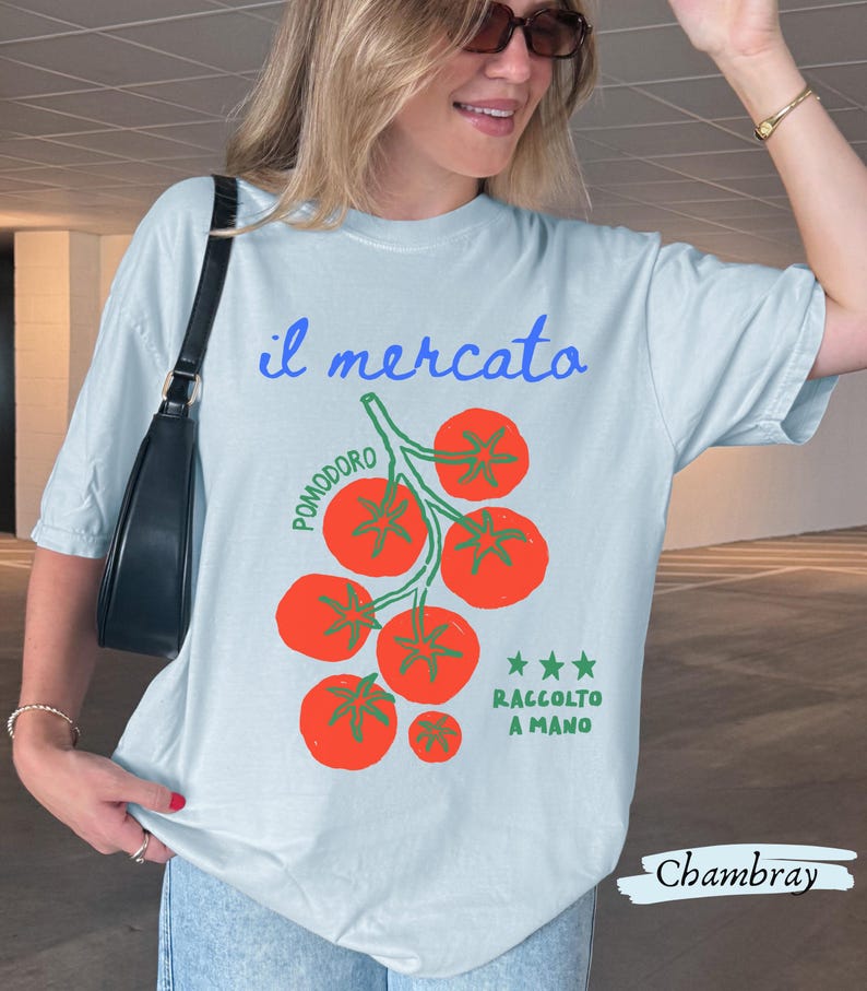 May include: Light blue t-shirt with the Italian text "il mercato" and a graphic of red tomatoes on a green vine. The text "Pomodoro" and "Raccolto a Mano" are also present. The word "Chambray" is written at the bottom.