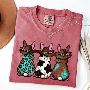 May include: A dusty rose Comfort Colors t-shirt featuring three cartoon bunnies wearing cowboy hats. The bunnies have different patterns: leopard print, cow print, and a brown and turquoise design. A silver necklace with a pendant is also visible.