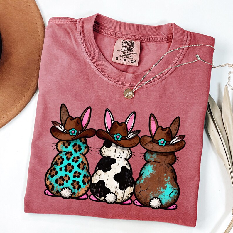 May include: A dusty rose Comfort Colors t-shirt featuring three cartoon bunnies wearing cowboy hats. The bunnies have different patterns: leopard print, cow print, and a brown and turquoise design. A silver necklace with a pendant is also visible.