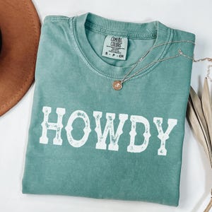 May include: A seafoam green t-shirt with the word "HOWDY" in distressed white lettering. A gold-coloured necklace with a pendant rests on the shirt. The shirt is a Comfort Colors brand, size S-CH. A brown hat is partially visible.
