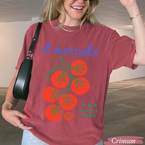 May include: Crimson-colored t-shirt featuring a tomato illustration with the text "il mercato" in blue. The design includes a cluster of red tomatoes with green stems and the text "RACCOLTO A MANO" in green. The shirt is a casual, comfortable style.