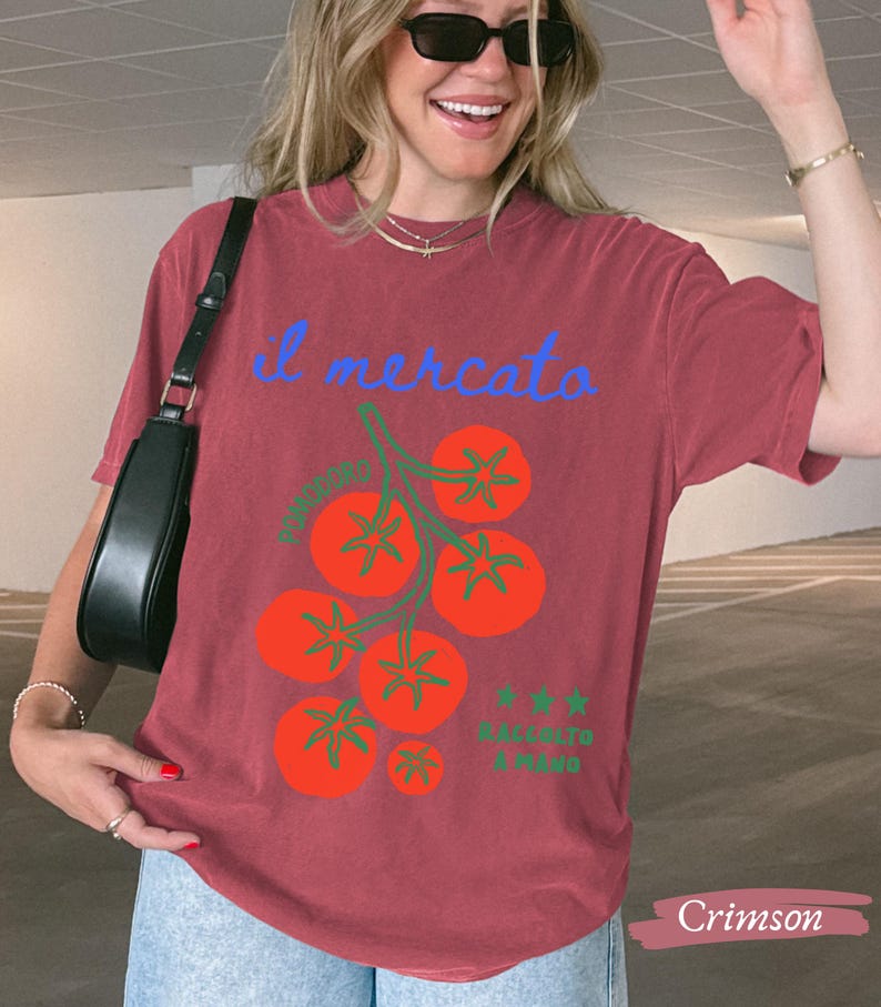 May include: Crimson-colored t-shirt featuring a tomato illustration with the text "il mercato" in blue. The design includes a cluster of red tomatoes with green stems and the text "RACCOLTO A MANO" in green. The shirt is a casual, comfortable style.