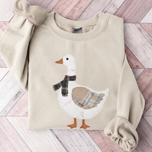 May include: Beige sweatshirt featuring an appliqued goose design. The goose has a white body, brown feet, and a plaid scarf. The sweatshirt has long sleeves with rolled cuffs, and is folded on a wooden surface.