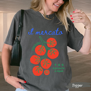 May include: Pepper-colored t-shirt with a graphic of red tomatoes on a green vine. The text "il mercato" is in blue, with "Pomodoro" and "Raccolto a Mano" also on the shirt. The shirt is a relaxed fit.
