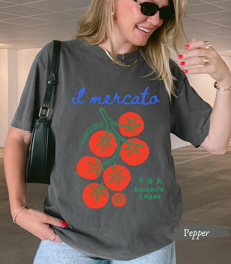 May include: Pepper-colored t-shirt with a graphic of red tomatoes on a green vine. The text "il mercato" is in blue, with "Pomodoro" and "Raccolto a Mano" also on the shirt. The shirt is a relaxed fit.