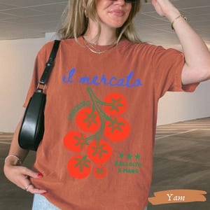May include: A rust-colored t-shirt with the text "il mercato" in blue script above a graphic of red tomatoes on green stems. The text "POMODORO" and "RACCOLTO A MANO" are also visible. The shirt is a casual style.