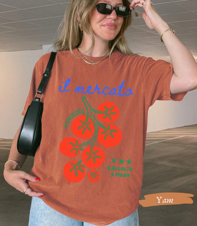 May include: A rust-colored t-shirt with the text "il mercato" in blue script above a graphic of red tomatoes on green stems. The text "POMODORO" and "RACCOLTO A MANO" are also visible. The shirt is a casual style.