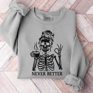 May include: A pale grey sweatshirt with a black graphic of a skeleton holding a coffee cup and giving an "OK" sign. The words "NEVER BETTER" are printed beneath the skeleton. The sweatshirt has long sleeves and a crew neck.