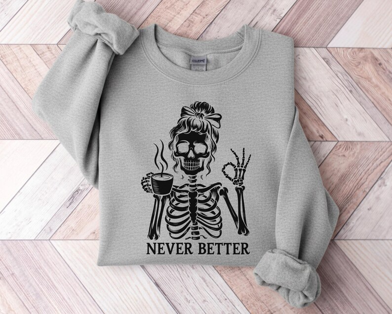 May include: A pale grey sweatshirt with a black graphic of a skeleton holding a coffee cup and giving an "OK" sign. The words "NEVER BETTER" are printed beneath the skeleton. The sweatshirt has long sleeves and a crew neck.