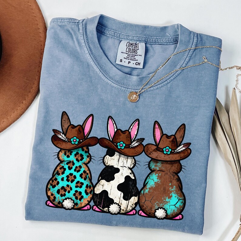 May include: A light blue Comfort Colors t-shirt featuring three cartoon bunnies with cowboy hats and turquoise accents. The bunnies have different patterns: leopard print, black and white cow spots, and brown with turquoise. A gold necklace is draped across the shirt.