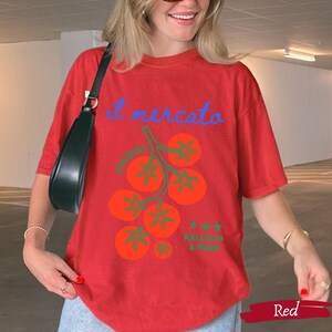 May include: Red t-shirt with a tomato graphic and the words "il mercato" in blue script. The graphic includes orange tomatoes and green stems. The text "Raccoglie a mano" is also present. The shirt is a casual, oversized fit.
