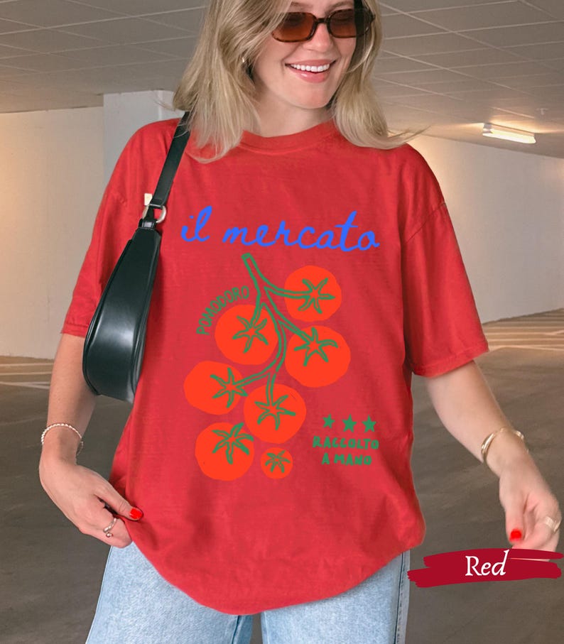 May include: Red t-shirt with a tomato graphic and the words "il mercato" in blue script. The graphic includes orange tomatoes and green stems. The text "Raccoglie a mano" is also present. The shirt is a casual, oversized fit.