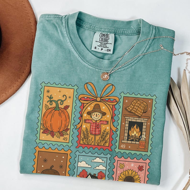Puede incluir: Una camiseta Comfort Colors turquesa con un estampado de sellos postales con tem&aacute;tica oto&ntilde;al. Los sellos representan una calabaza, un espantap&aacute;jaros, una chimenea y otras im&aacute;genes de oto&ntilde;o. Un collar plateado con un colgante descansa sobre la camiseta.