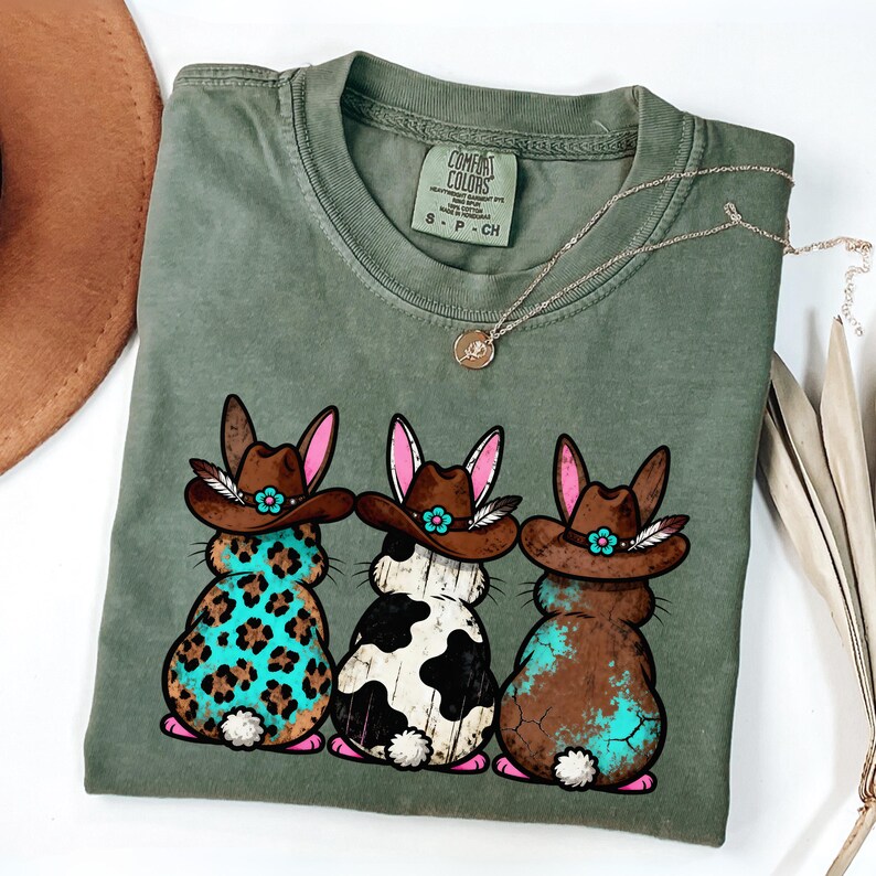 May include: Sage green Comfort Colors t-shirt featuring three cartoon bunnies wearing cowboy hats. The bunnies have different patterns: leopard print, black and white cow print, and brown with turquoise accents. A silver necklace with a pendant is also visible.