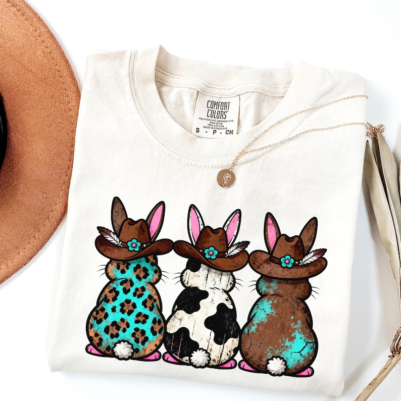 May include: Cream-colored t-shirt featuring three cartoon bunnies wearing cowboy hats. Each bunny has a different pattern: leopard print, cow print, and a distressed turquoise design. The shirt has a Comfort Colors tag and a gold necklace.