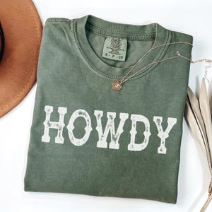May include: An olive green t-shirt with the word "HOWDY" in distressed white lettering. A gold necklace with a pendant rests on the shirt. A brown hat is partially visible in the upper left corner. The shirt has a Comfort Colors tag.
