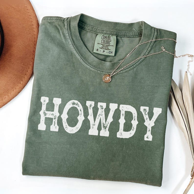 May include: An olive green t-shirt with the word "HOWDY" in distressed white lettering. A gold necklace with a pendant rests on the shirt. A brown hat is partially visible in the upper left corner. The shirt has a Comfort Colors tag.