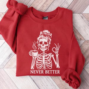 May include: A red sweatshirt featuring a white skeleton design. The skeleton is holding a coffee cup and giving the okay sign. The text "NEVER BETTER" is below the graphic.