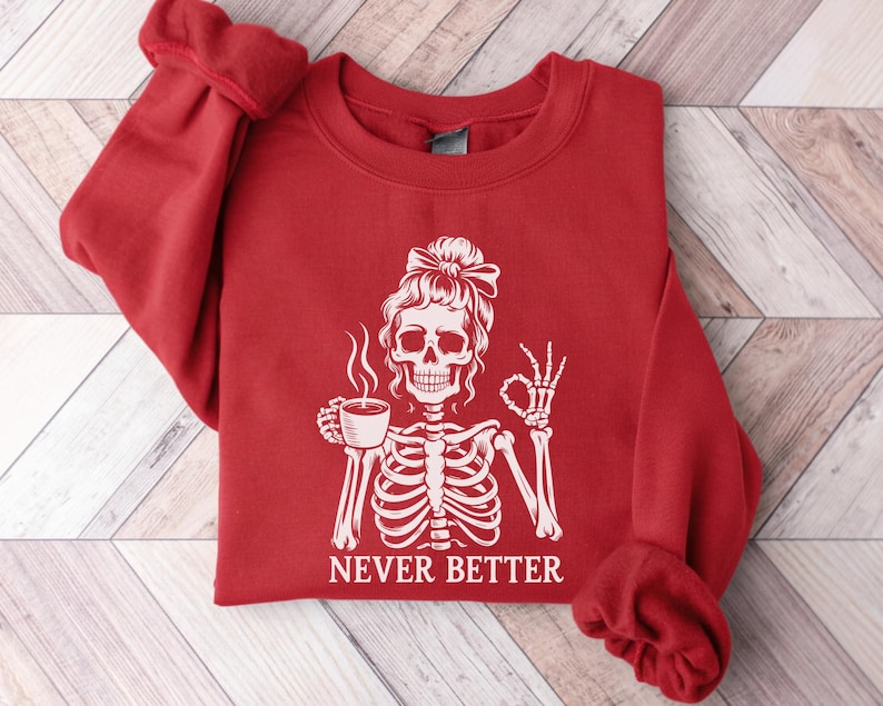 May include: A red sweatshirt featuring a white skeleton design. The skeleton is holding a coffee cup and giving the okay sign. The text "NEVER BETTER" is below the graphic.