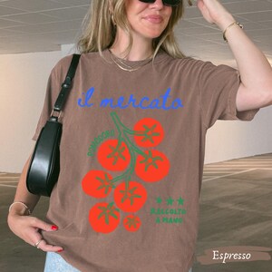 May include: Espresso-colored t-shirt with a tomato illustration. The design features a cluster of red tomatoes with green stems and the text "il mercato" in blue. Additional text includes "Pomodoro" and "Raccolto a Mano."