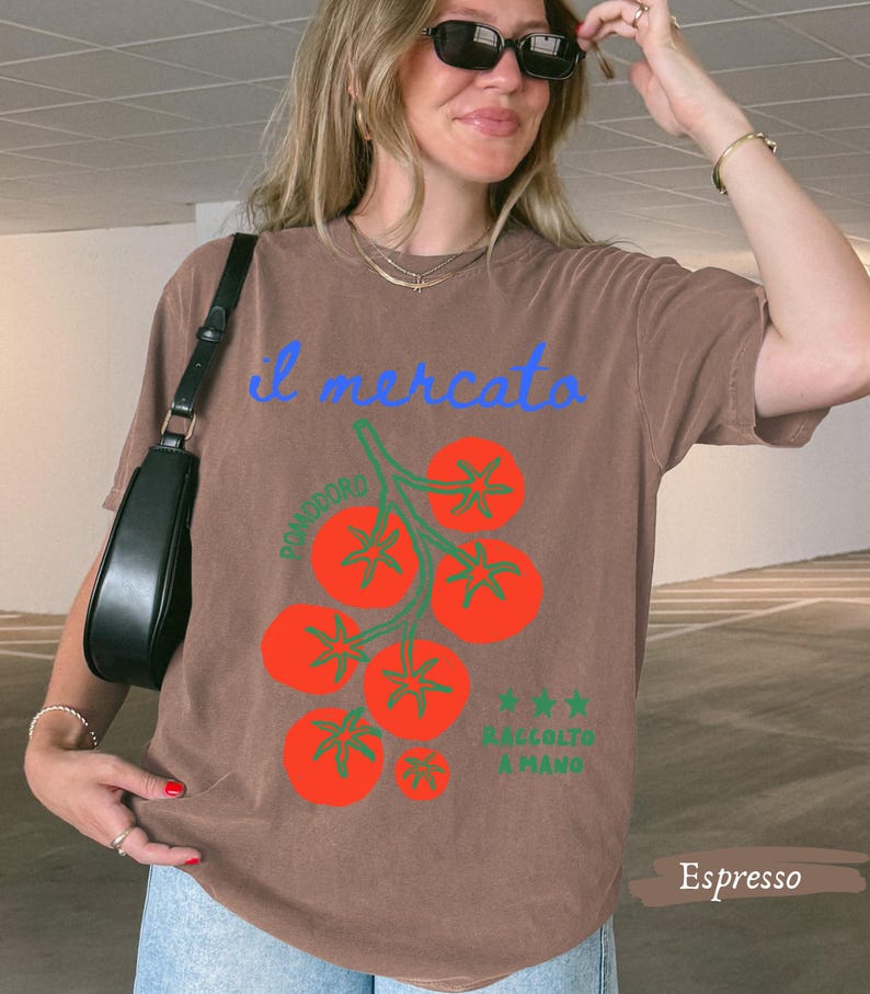 May include: Espresso-colored t-shirt with a tomato illustration. The design features a cluster of red tomatoes with green stems and the text "il mercato" in blue. Additional text includes "Pomodoro" and "Raccolto a Mano."