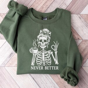 May include: A dark green sweatshirt featuring a white skeleton graphic holding a coffee cup and giving an "okay" sign. The words "NEVER BETTER" are printed below. The sweatshirt has long sleeves and a crew neck.