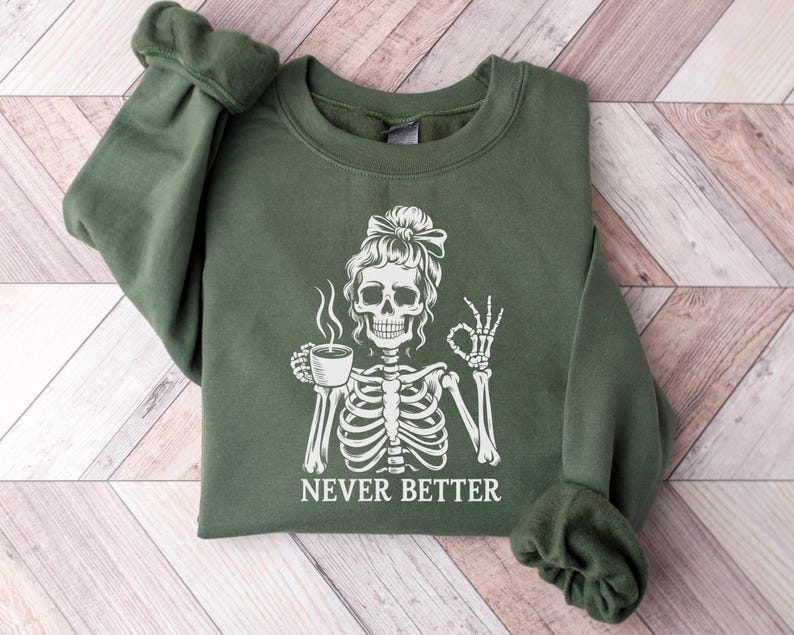 May include: A dark green sweatshirt featuring a white skeleton graphic holding a coffee cup and giving an "okay" sign. The words "NEVER BETTER" are printed below. The sweatshirt has long sleeves and a crew neck.