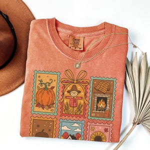 Puede incluir: Camiseta naranja Comfort Colors con un dise&ntilde;o de sellos coloridos con ilustraciones de tem&aacute;tica oto&ntilde;al, que incluyen una calabaza, un espantap&aacute;jaros y una chimenea. La camiseta tiene cuello redondo y mangas cortas. Tambi&eacute;n se ve un collar dorado y un sombrero marr&oacute;n.