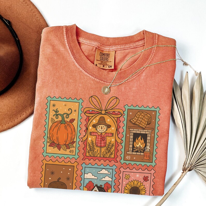 Puede incluir: Camiseta naranja Comfort Colors con un dise&ntilde;o de sellos coloridos con ilustraciones de tem&aacute;tica oto&ntilde;al, que incluyen una calabaza, un espantap&aacute;jaros y una chimenea. La camiseta tiene cuello redondo y mangas cortas. Tambi&eacute;n se ve un collar dorado y un sombrero marr&oacute;n.