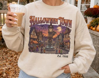 May include: A beige crewneck sweatshirt featuring a graphic print of a town square at night with the text HALLOWEEN TOWN and Est. 1998. The illustration depicts buildings, a clock tower, a fountain, and figures under a full moon and bats.