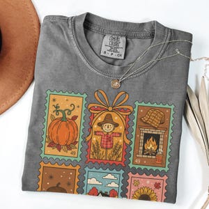 Puede incluir: Camiseta gris Comfort Colors con un gr&aacute;fico colorido de sellos postales con tem&aacute;tica oto&ntilde;al. Los sellos representan una calabaza, un espantap&aacute;jaros, una chimenea y un girasol. Un collar plateado con un colgante est&aacute; sobre la camiseta.