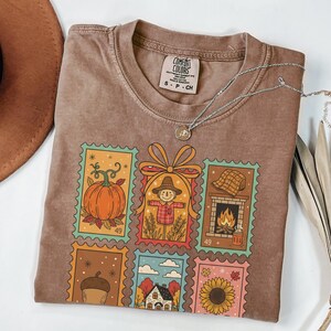 Puede incluir: Camiseta Comfort Colors marr&oacute;n con un dise&ntilde;o de sellos con tem&aacute;tica oto&ntilde;al. Los sellos presentan una calabaza, un espantap&aacute;jaros, una chimenea, una bellota, una casa y un girasol. Un collar plateado con un colgante est&aacute; sobre la camiseta.