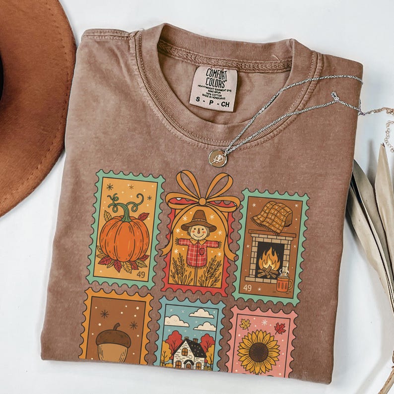 Puede incluir: Camiseta Comfort Colors marr&oacute;n con un dise&ntilde;o de sellos con tem&aacute;tica oto&ntilde;al. Los sellos presentan una calabaza, un espantap&aacute;jaros, una chimenea, una bellota, una casa y un girasol. Un collar plateado con un colgante est&aacute; sobre la camiseta.