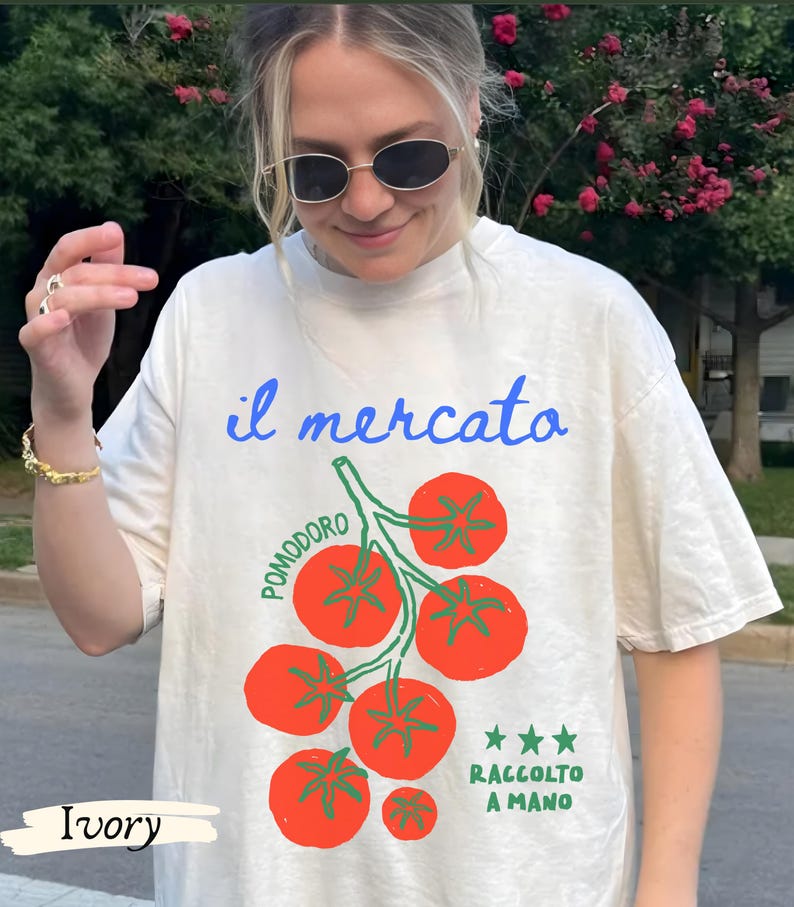 May include: Ivory-colored t-shirt with a graphic of red tomatoes on a green vine. The text "il mercato" is in blue script above the tomatoes, with "Pomodoro" and "Raccolto a Mano" in green. The shirt is a casual, relaxed fit.