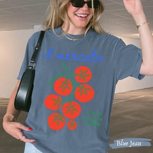 May include: Blue jean colored t-shirt with the text "il mercato" in light blue script. The shirt features a graphic of red tomatoes with green stems. The words "il mercato" are above the tomato graphic.
