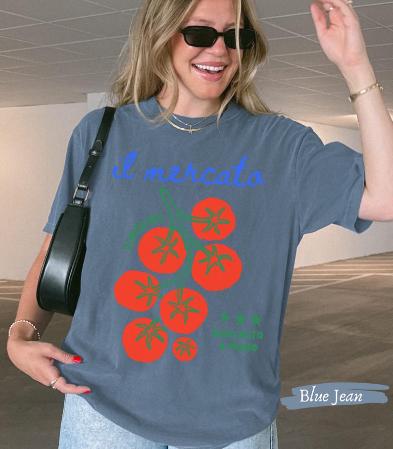 May include: Blue jean colored t-shirt with the text "il mercato" in light blue script. The shirt features a graphic of red tomatoes with green stems. The words "il mercato" are above the tomato graphic.