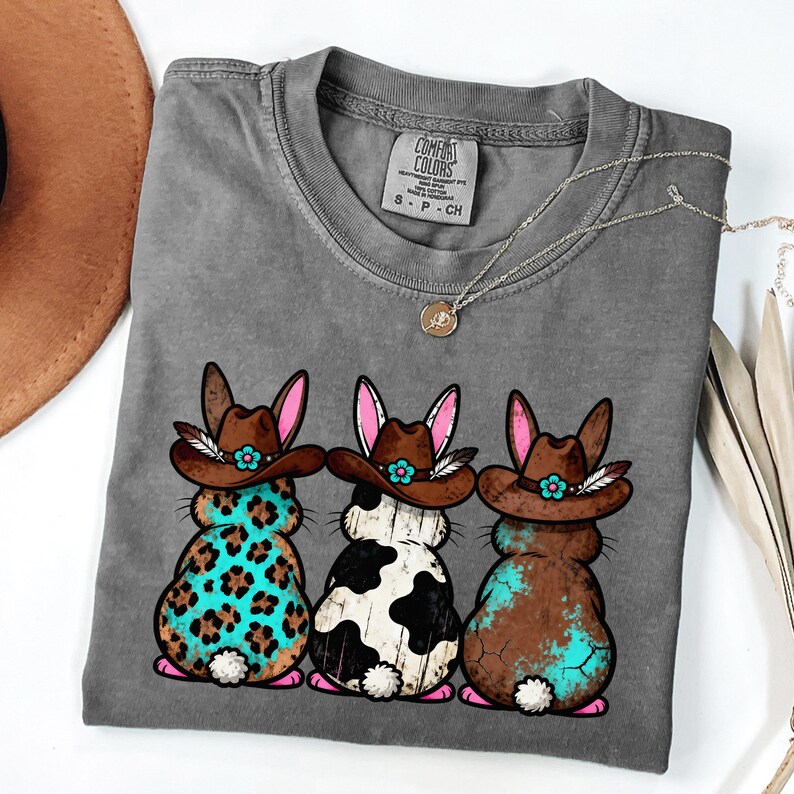 May include: Gray Comfort Colors t-shirt featuring three cartoon bunnies wearing cowboy hats. The bunnies have different patterns: leopard print, black and white spots, and brown with turquoise accents. A silver necklace with a pendant is also visible.