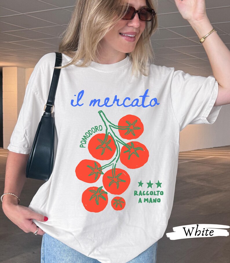 May include: White t-shirt with a tomato graphic. The design includes the Italian words "il mercato" and "pomodoro," along with "raccolto a mano." The graphic features red tomatoes and green stems. The word "White" is printed at the bottom.