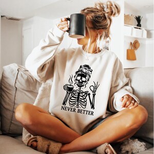 May include: A cream-coloured sweatshirt featuring a black skeleton graphic holding a coffee cup and the 'okay' hand gesture. The text "NEVER BETTER" is below the graphic. A black mug is held in the image.