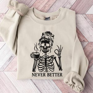 May include: Cream-coloured sweatshirt featuring a black skeleton design. The skeleton holds a coffee cup and gives an 'okay' sign. The words "NEVER BETTER" are printed beneath. The sweatshirt has a crew neck and long sleeves.
