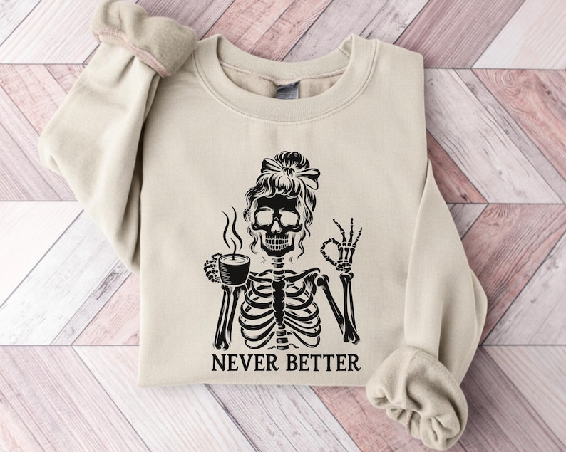 May include: Cream-coloured sweatshirt featuring a black skeleton design. The skeleton holds a coffee cup and gives an 'okay' sign. The words "NEVER BETTER" are printed beneath. The sweatshirt has a crew neck and long sleeves.