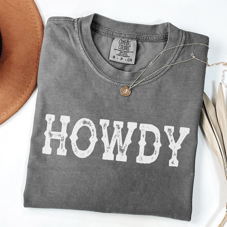 May include: A grey Comfort Colors t-shirt with the word "HOWDY" in white, distressed lettering. A gold pendant necklace and a brown hat are also visible. The shirt is folded and laid flat on a white surface.