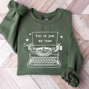 May include: An olive green sweatshirt with the phrase "You're just my type" and a white illustration of a typewriter. Two small white hearts are on the top right and left of the typewriter.