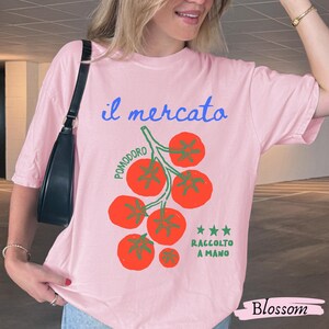 May include: A light pink t-shirt with the Italian text "il mercato" in blue script and a graphic of red tomatoes on a green vine. The text "POMODORO" and "RACCOLTO A MANO" are also present. The word "Blossom" is written in a cursive font.