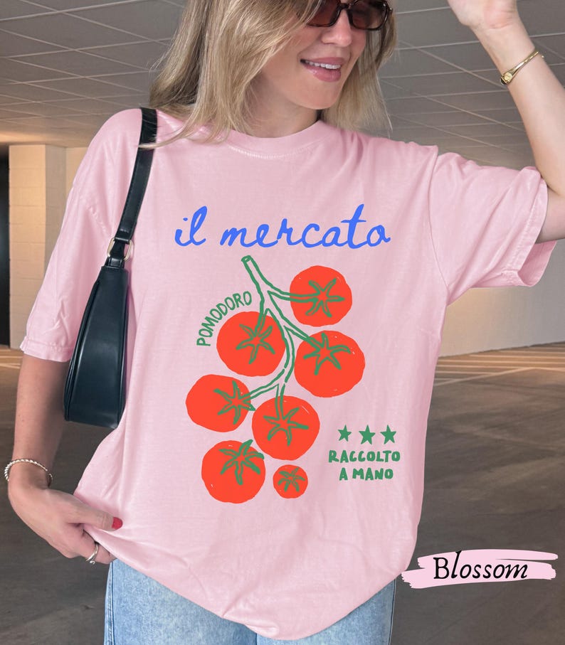 May include: A light pink t-shirt with the Italian text "il mercato" in blue script and a graphic of red tomatoes on a green vine. The text "POMODORO" and "RACCOLTO A MANO" are also present. The word "Blossom" is written in a cursive font.