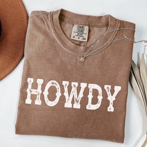 May include: A brown Comfort Colors t-shirt with the word "HOWDY" in white block letters. A silver necklace with a small pendant rests on the shirt. A brown hat is visible in the upper left corner.