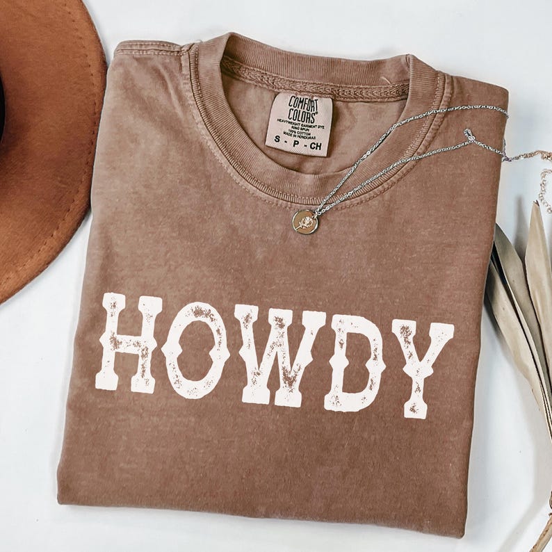 May include: A brown Comfort Colors t-shirt with the word "HOWDY" in white block letters. A silver necklace with a small pendant rests on the shirt. A brown hat is visible in the upper left corner.