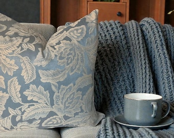 May include: A decorative throw pillow with a blue background and a cream-colored floral pattern. A blue knitted throw blanket and a blue teacup and saucer are on a gray sofa. The scene is set in a cozy living room.