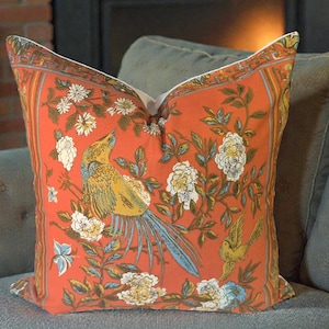 Peut inclure: Un coussin décoratif orange avec un motif d'oiseau et de fleurs. Le coussin présente une illustration détaillée d'un oiseau aux plumes jaunes, bleues et vertes, entouré de fleurs blanches et bleues et de feuilles vertes. Le coussin est posé sur un canapé gris.