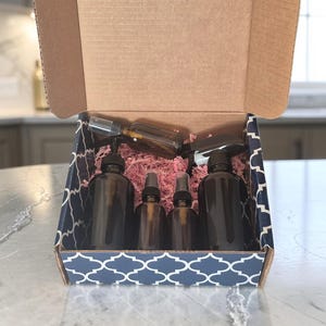Peut inclure: Une boîte d'expédition en carton avec un motif quadrilobé bleu marine et blanc contenant plusieurs flacons en verre ambré foncé avec des pompes et des vaporisateurs noirs, nichés dans du papier déchiqueté rose pour la protection.